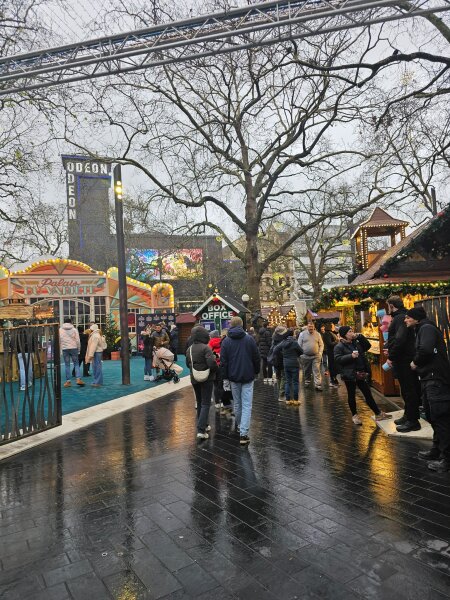 Christmas market on Leicester Square with wooden chalets and a fairground ride on wet paving.