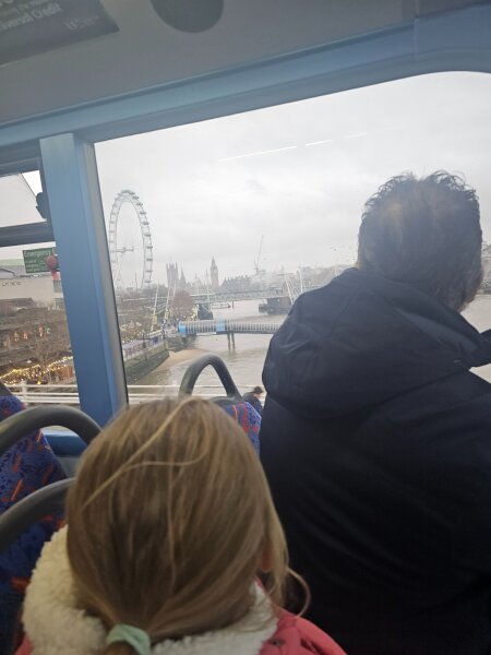 View from the top deck of a double-decker bus across the Thames, with the London Eye and Big Ben in the haze and a child on the front seat.