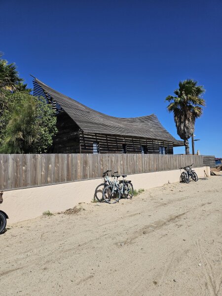 Houten strandpaviljoen met een gebogen dak van latten, met fietsen tegen een muurtje langs het zandpad ernaartoe