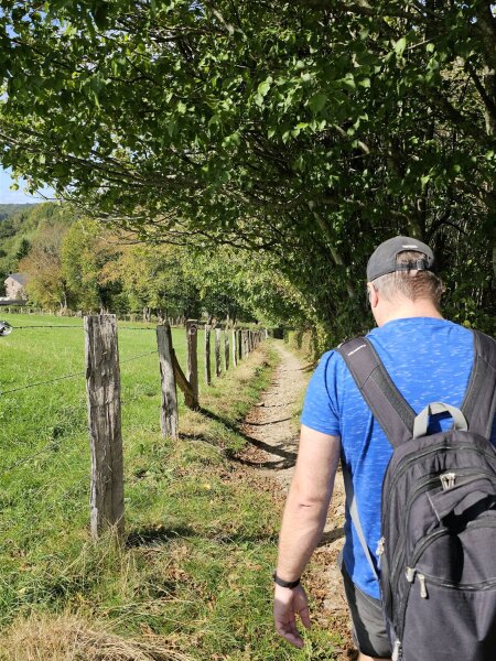 Man with a backpack walking a narrow dirt path along a meadow with wooden fence posts