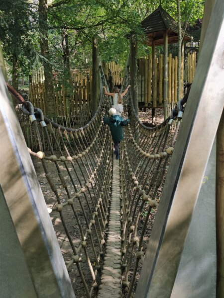 Two children seen from behind crossing a rope bridge to a wooden play tower in the woods