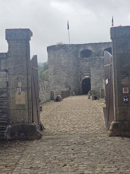 Open wooden gate and a cobbled path leading to the stone tower of Bouillon castle under a grey sky